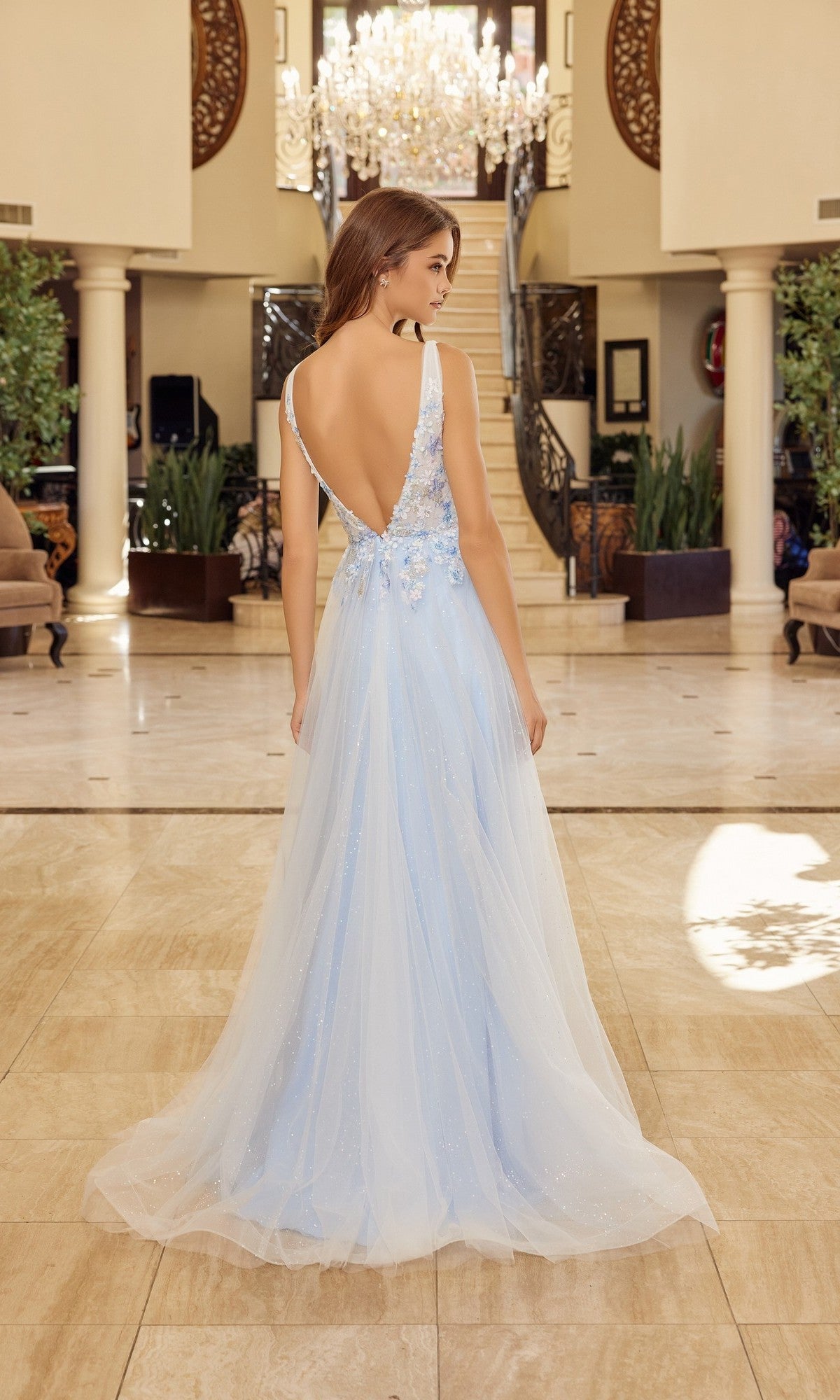 A woman in the Nox Anabel Y1799 long prom dress, featuring pastel tulle, a deep V-back, and floral details, stands in an elegant interior with a grand staircase and chandelier.